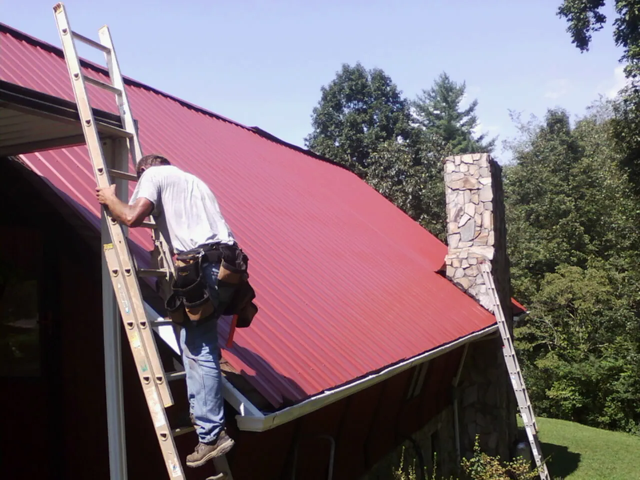 Roofer on a ladder beside a red metal roof and stone chimney — Soffit & Fascia Repair by Roofing Brothers in North Scituate